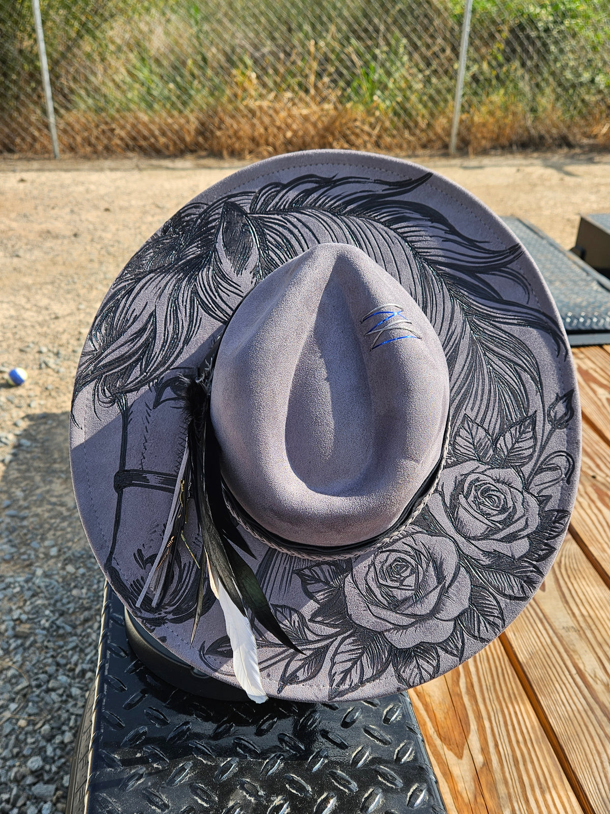 A wide-brimmed felt hat in a muted lavender-gray color, decorated with black line art illustrations of a horse and roses. The hat features a matching fabric band and a white feather accent, resting on a black diamond-plate metal surface outdoors.