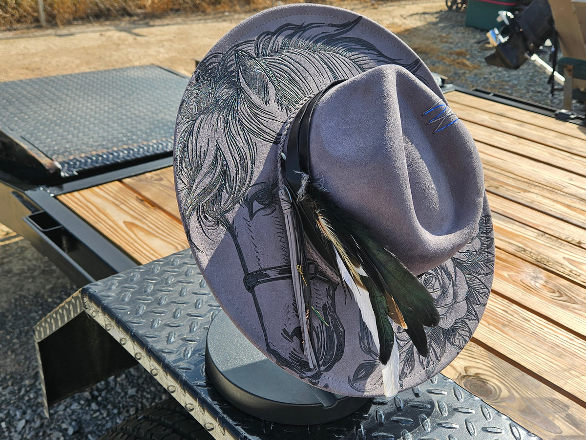 A wide-brimmed hat with a black and white illustrated pattern on a light gray base, featuring a matching gray crown and dark black and white feathers, displayed on a metal stand on a wooden trailer.