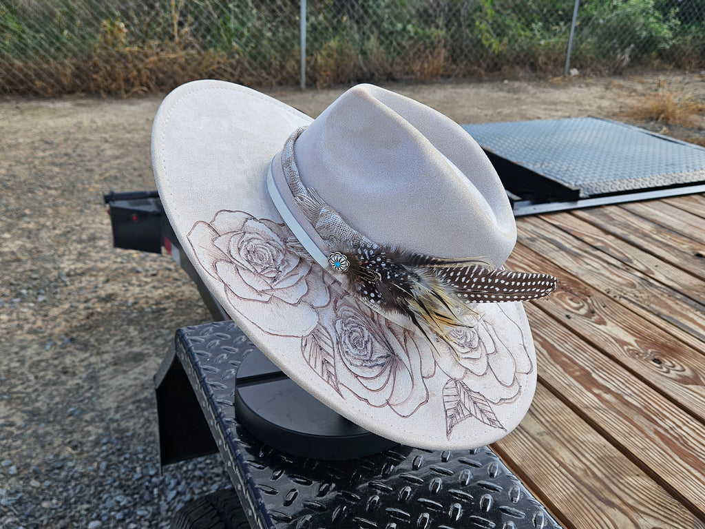 A light beige wide-brim fedora hat with embroidered rose patterns on the brim, a matching band, and a feather accent, displayed on a black metal stand outdoors on a wooden deck.
