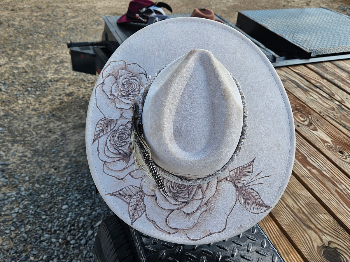 A light beige cowboy hat with engraved rose designs on the brim, resting on a metal trailer platform outdoors.