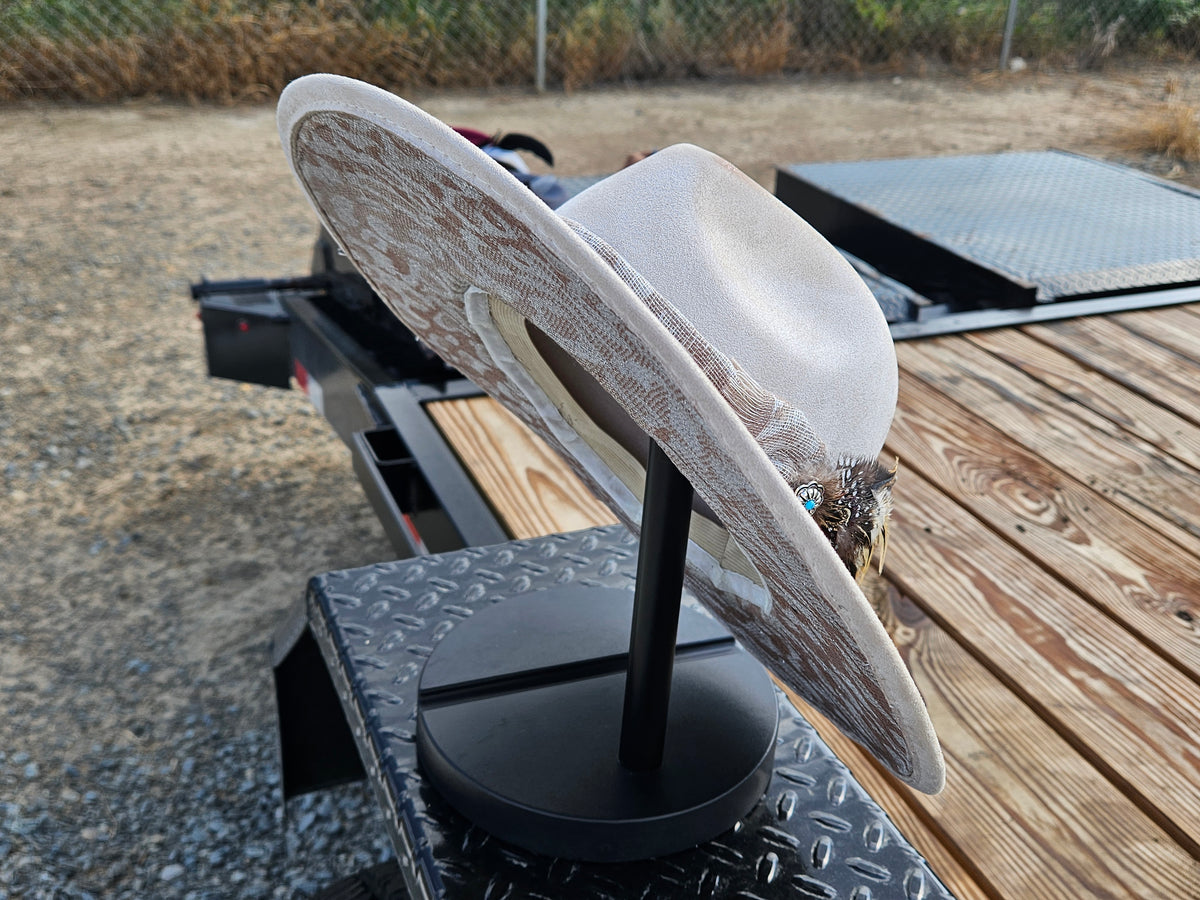 A light beige western-style hat with a brown textured pattern and a small turquoise accent, displayed on a black stand on a trailer deck outdoors.