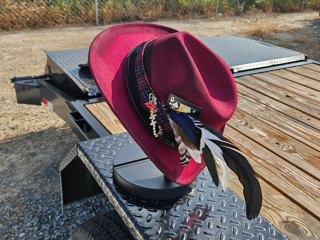 A burgundy felt cowboy hat with a black patterned band, decorative beads, and black, white, and gray feathers, displayed on a black metal trailer ramp outdoors.