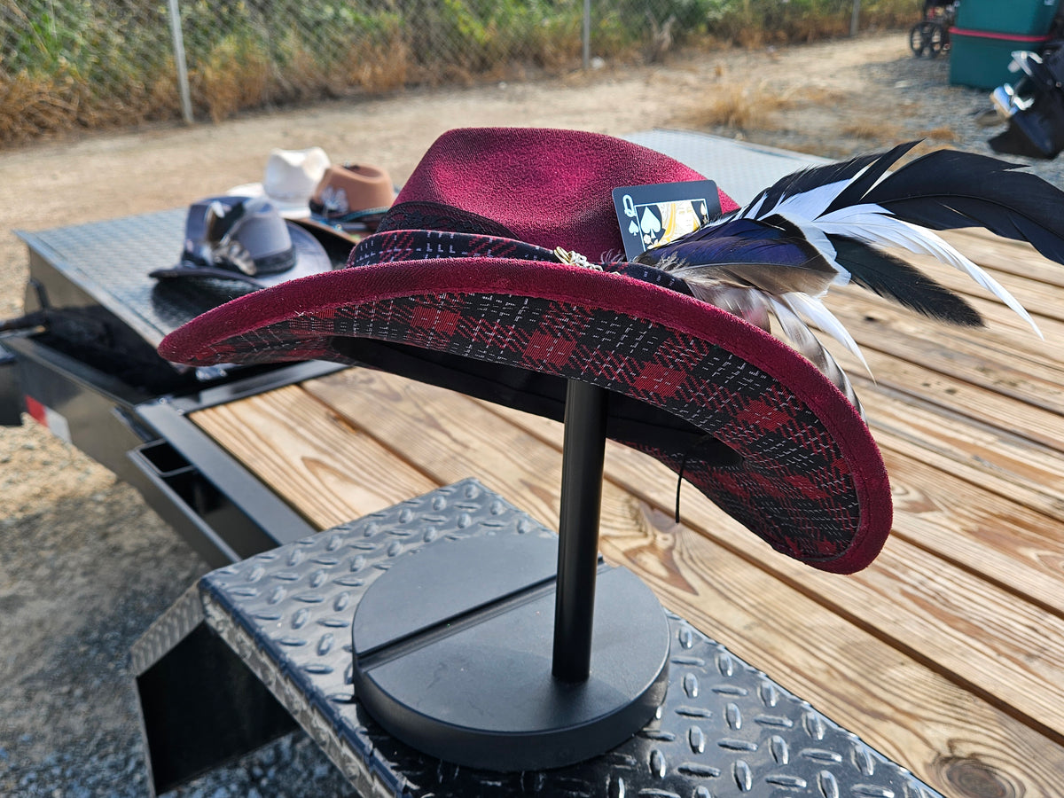A wide-brimmed hat displayed on a black stand outdoors on a wooden trailer. The hat is burgundy with a burgundy and black plaid brim and is decorated with black, white, and blue feathers.