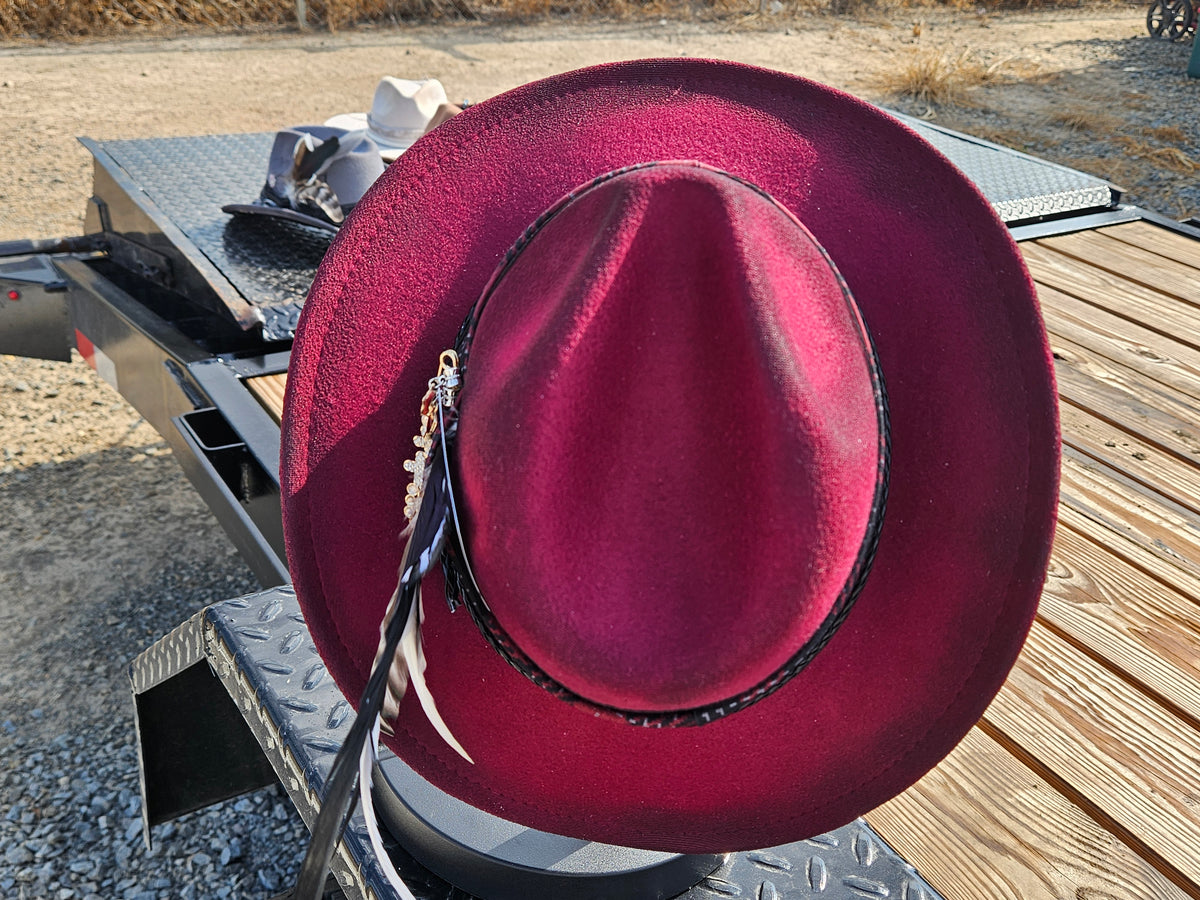A wide-brimmed western-style hat in deep burgundy red, featuring a black band with silver-toned studs and a hanging feather ornament, displayed on a metal trailer in an outdoor setting.