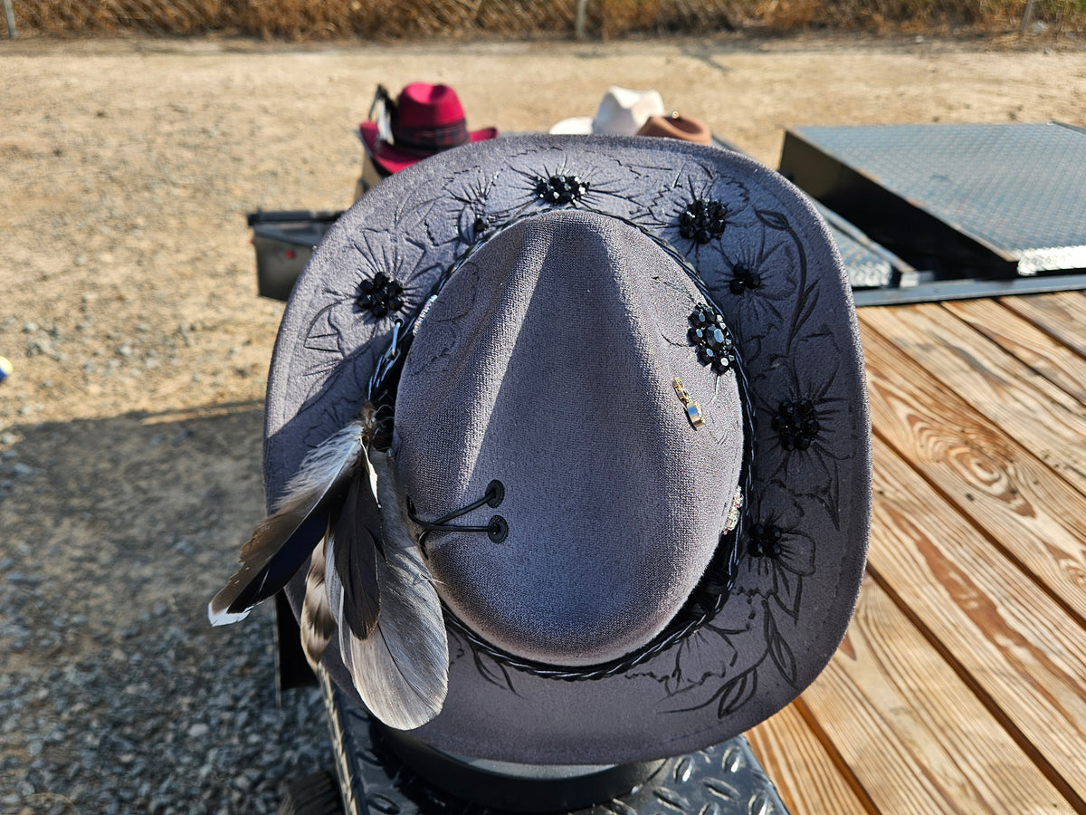 A wide-brimmed gray cowboy hat with black floral embroidery and feather accents, photographed outdoors on a wooden deck with gravel and another red hat in the background.