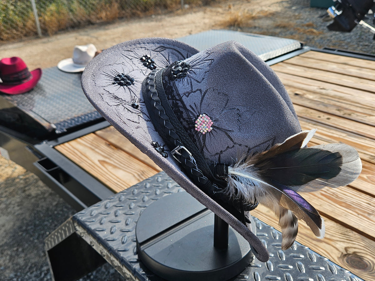 A dark gray felt cowboy hat with black floral embroidery, a braided black band, a pink rhinestone center, and brown, black, and purple feathers, displayed on a black stand on a wooden deck.
