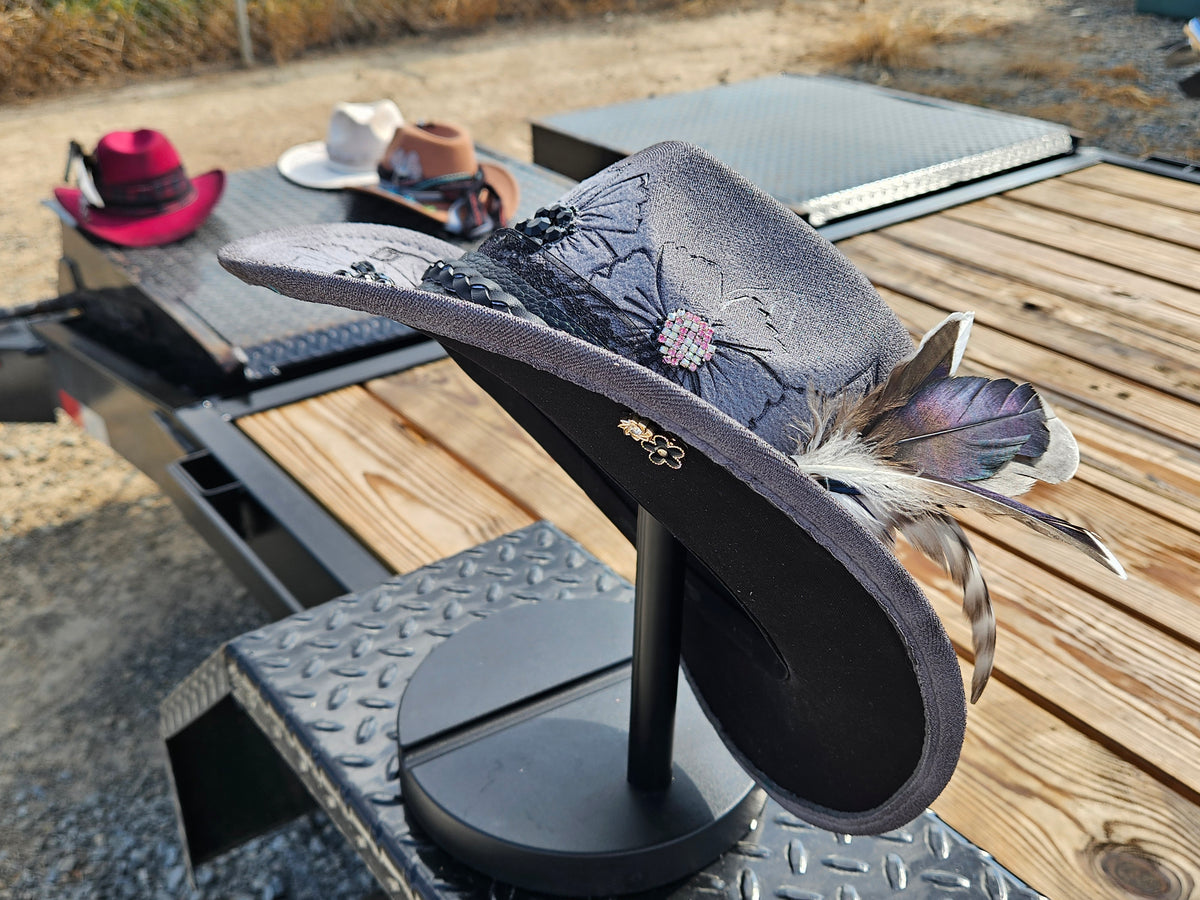 A dark gray felt cowboy hat with black floral embroidery, a rhinestone flower, and feather accents, displayed on a black stand on a truck bed next to other hats.